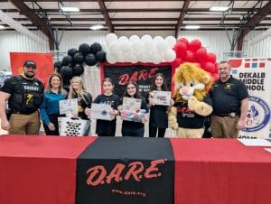 Sixth graders at DeKalb Middle School graduated from the D.A.R.E. (Drug Abuse Resistance Education) program in a ceremony Friday. Pictured from left are DMS School Resource Officer and D.A.R.E. Instructor Joseph Carroll, DeKalb Circuit Court Clerk Susan Martin, 1st Place Art Winner – Ava Gillis, 2nd Place – Sidney Rigsby, 3rd Place – McKenzie Hendrixson, Honorable Mention – Miller Williams, Darren the D.A.R.E. Lion mascot, and Sheriff Patrick Ray. (Shawna Willingham Photo)